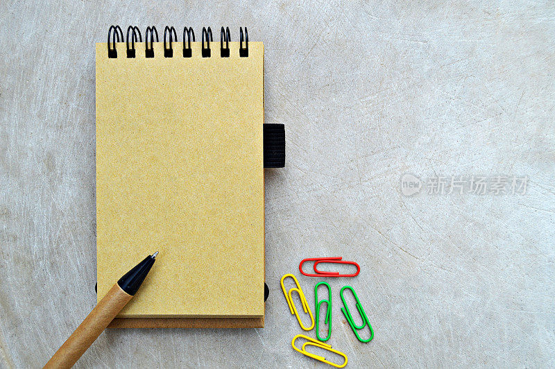 这是一张经典的水平照片，黄色的米黄色素色页，螺旋装订的垂直记事本，黑色和棕色的钢笔和彩色回形针放在木质的米黄色邋遢的背景上。
