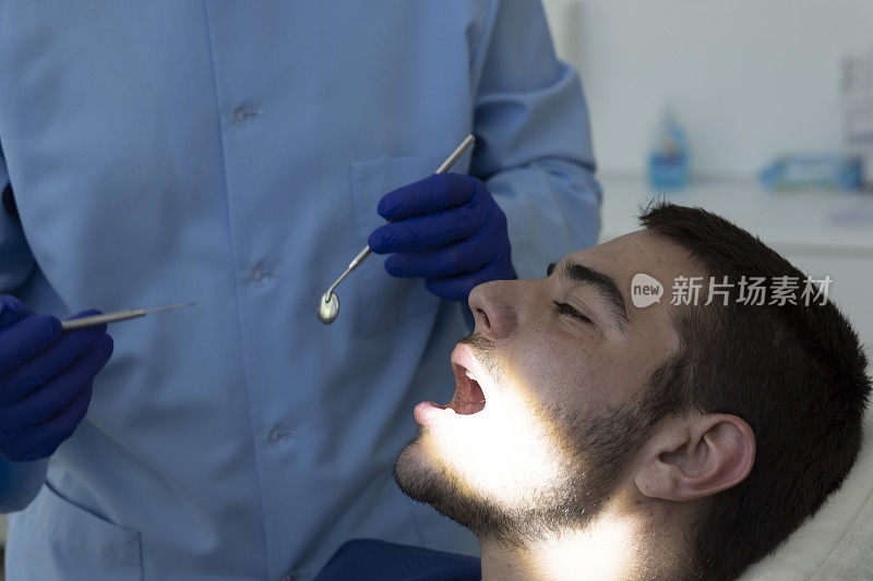 近距离拍摄专业牙医戴着防护口罩为病人做检查。一段视频显示，一名年轻男子坐在牙医的办公室里接受蛀牙治疗。