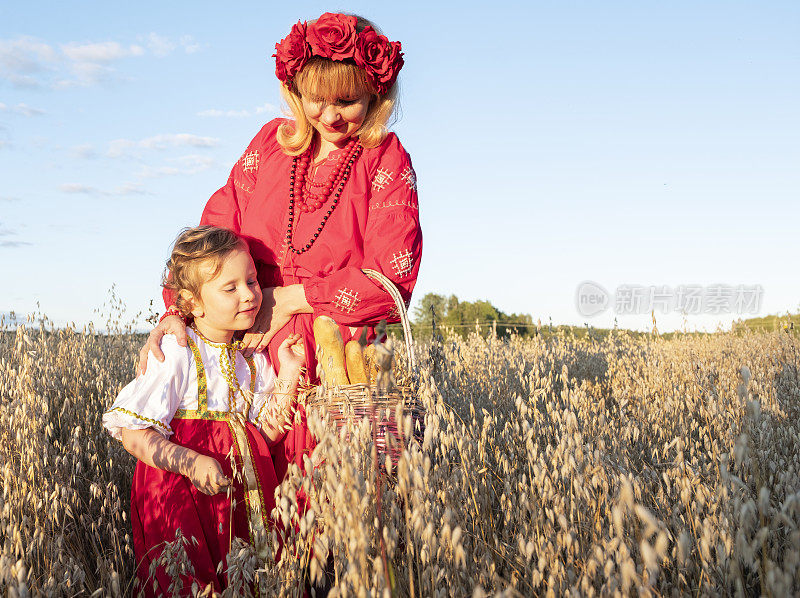 一个女孩拿着一个柳条篮子，里面放着新鲜的面包，背景是一片燕麦田。孩子，5岁女孩，拥抱妈妈。一个穿着红色衣服，头上戴着花环的女人。日落。