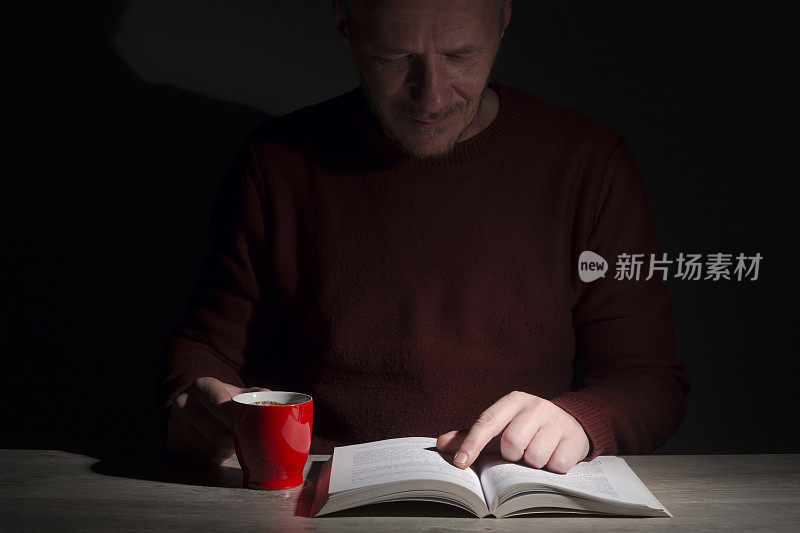 中年男子坐在书桌前，晚上在昏暗的房间里看书喝咖啡