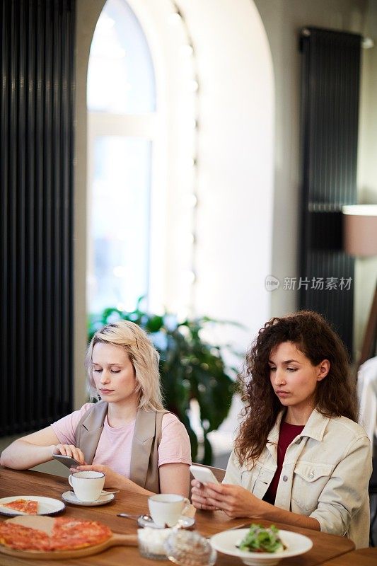 严肃的多民族女性朋友坐在餐桌边吃着菜，在现代咖啡馆里用着智能手机吃午餐，沟通不畅