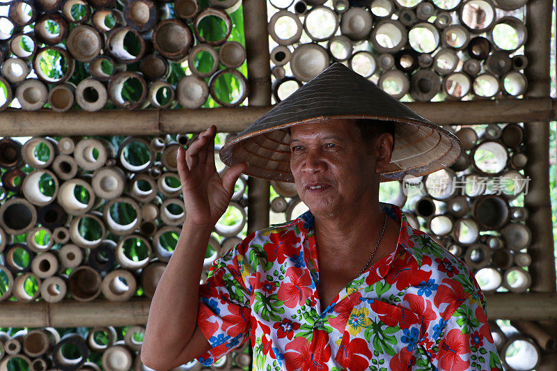 这名男子头戴越南圆锥形帽子，身穿五颜六色的花卉图案衬衫，站在竹屋的墙壁上。