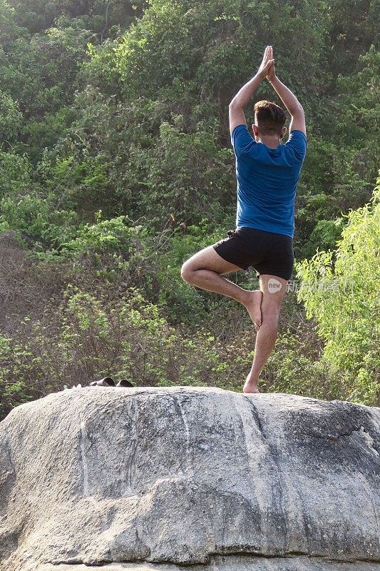 印度男子练习树姿势(Vrksasana)瑜伽位置在岩石上，后视图，水的边缘，帕洛伦海滩，果阿，印度