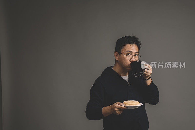 一个亚裔华人男子喝着一杯咖啡和一盘灰色背景的甜甜圈