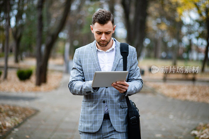 年轻帅气的经理一边在公园里散步一边在平板电脑上工作。