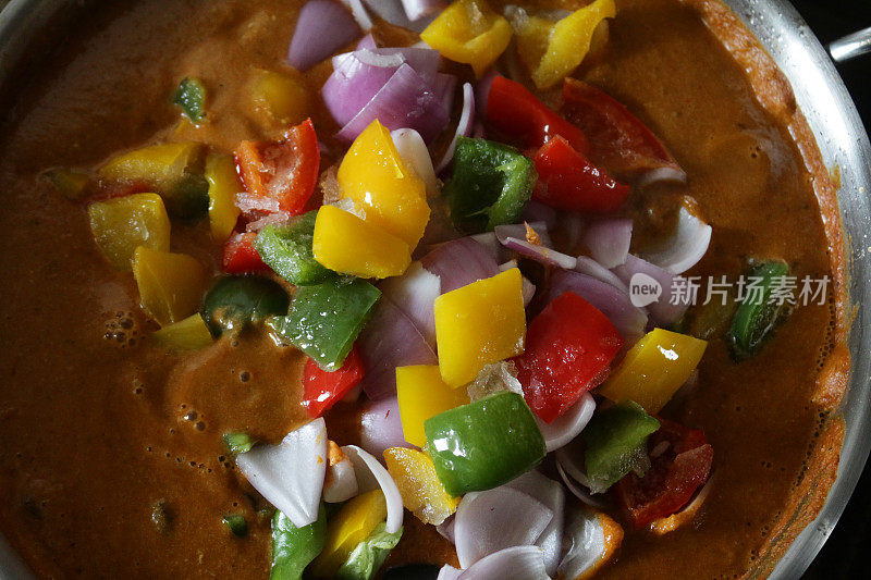 这是一个烹饪锅的特写图像，里面装满了鸡肉咖喱酱，冰冻的交通灯颜色，切碎的辣椒，红，绿，黄的甜椒和红洋葱