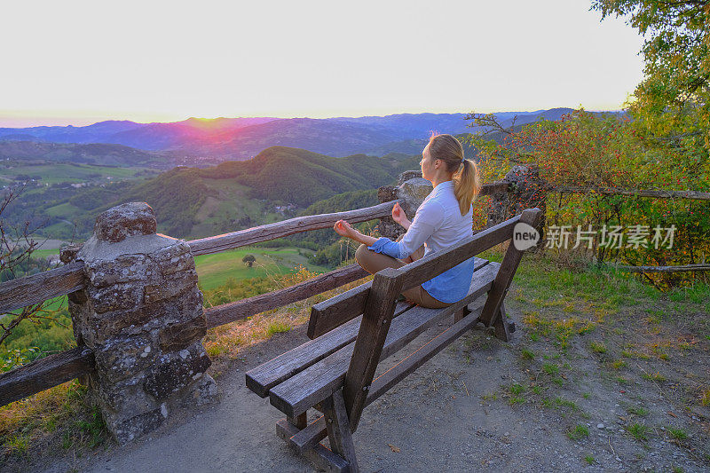 平静的白人女孩坐在长凳上，沉思在日落横跨山脉景观。放松，瑜伽，冷静，爱好概念。本空间