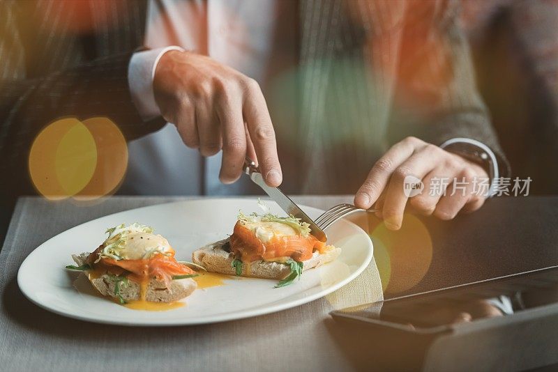 在一家室内餐厅里，一位年轻帅气的男士正在用他的手机打电话，同时享用美味的火腿蛋本尼迪克特与淡盐鲑鱼和酱汁