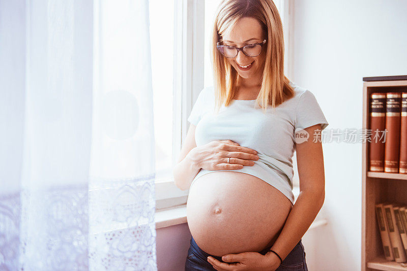一位年轻的孕妇抱着肚子站在窗边