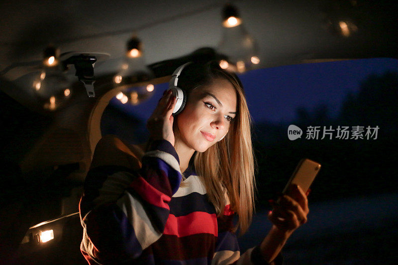一个年轻漂亮的女人露营在车的后面，一边放松一边用她的手机和耳机听音乐