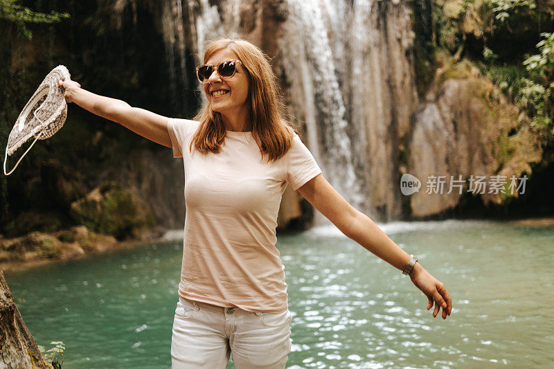 美丽的年轻女子手里拿着一顶帽子，站在瀑布旁边，探索大自然，呼吸新鲜空气