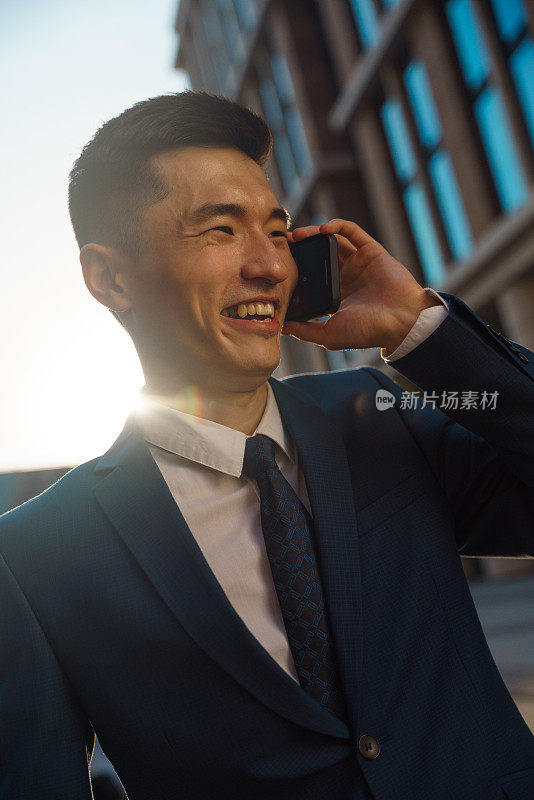 一个穿着西装的年轻亚洲英俊的商人正在听他朋友的电话，他笑了，他在商业区的一条街上