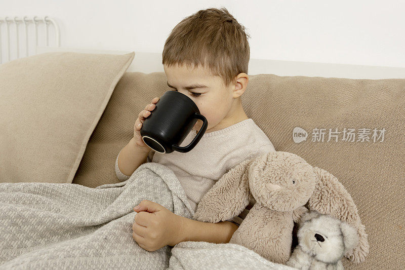 生病的小男孩在家里的床上喝热茶。生病的孩子用毯子裹着，房间里放着马克杯。流感季节。室内装饰和衣服采用自然的大地色。舒适的环境。