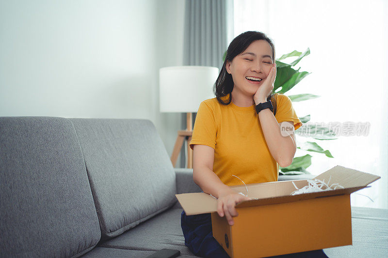 亚洲女人打开纸箱，开心兴奋地看着里面，坐在家里客厅的沙发上。