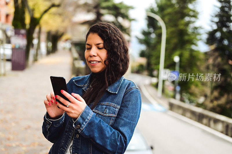 坦率开朗的中年成年女子牛仔夹克使用黑色智能手机户外。