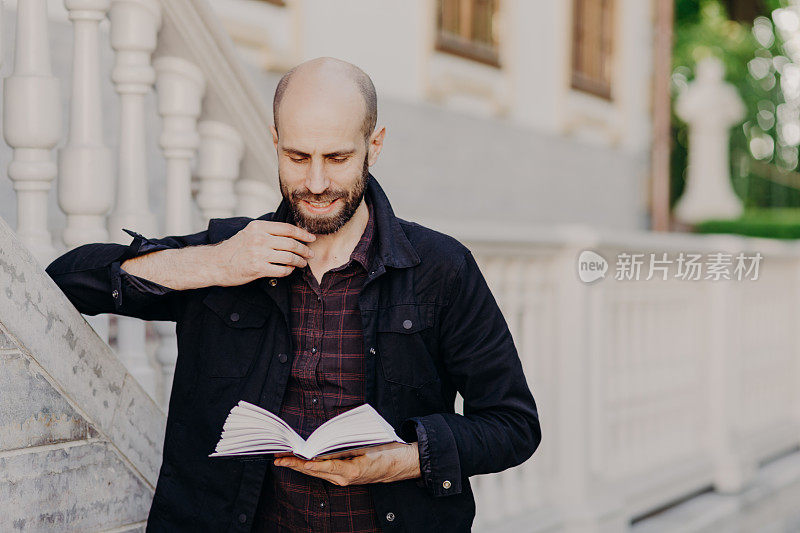 英俊伶俐的秃头男拿着书，用严肃的表情读有趣的小说，站在户外对着古老的白色建筑，喜欢读书。人，爱好，生活方式，休闲理念