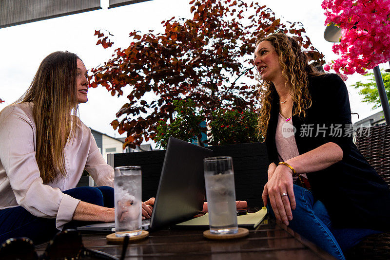 在室外咖啡馆的宁静氛围中，两个女人在点缀着层层叠叠的粉红色花朵的凉棚下相遇。一个女人穿着浅色的长袖上衣，而另一个留着长长的卷发，黑色夹克搭配粉红色衬衫。