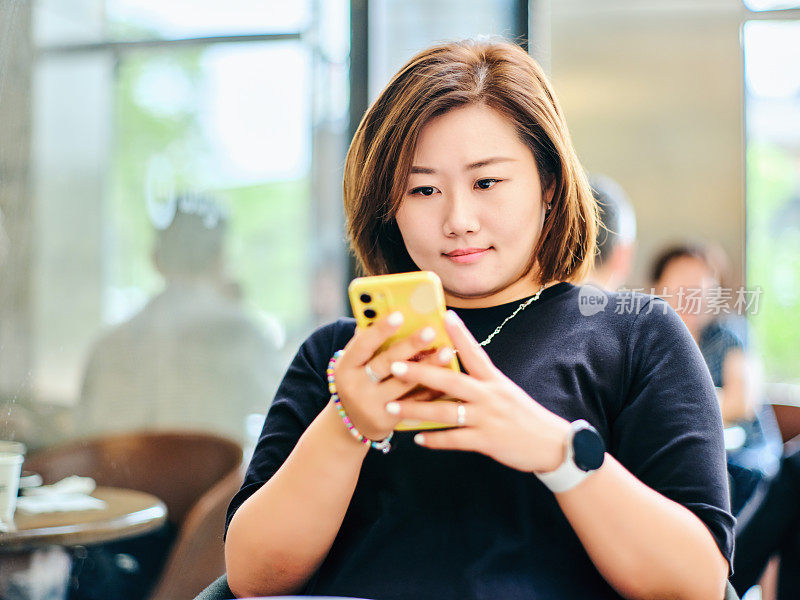 一位中国女士坐在咖啡店的窗边，用她的手机上网