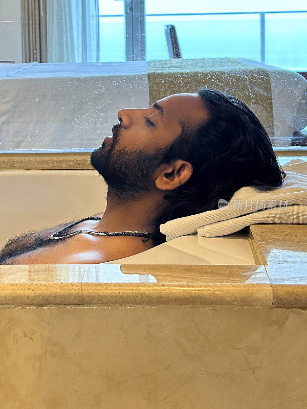 特写照片:印度男子躺在白色浴缸里，闭着眼睛，在浴缸里放松，头枕在浴盆边的毛巾上，毛茸茸的胸部，面部毛发，头像，侧面视图