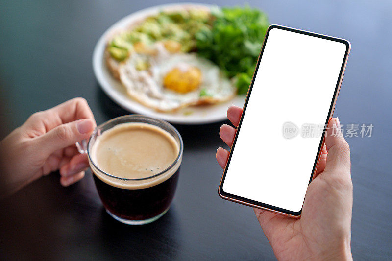 一个女人在家里吃早餐时拿着智能手机，智能手机显示空白屏幕作为设计模型