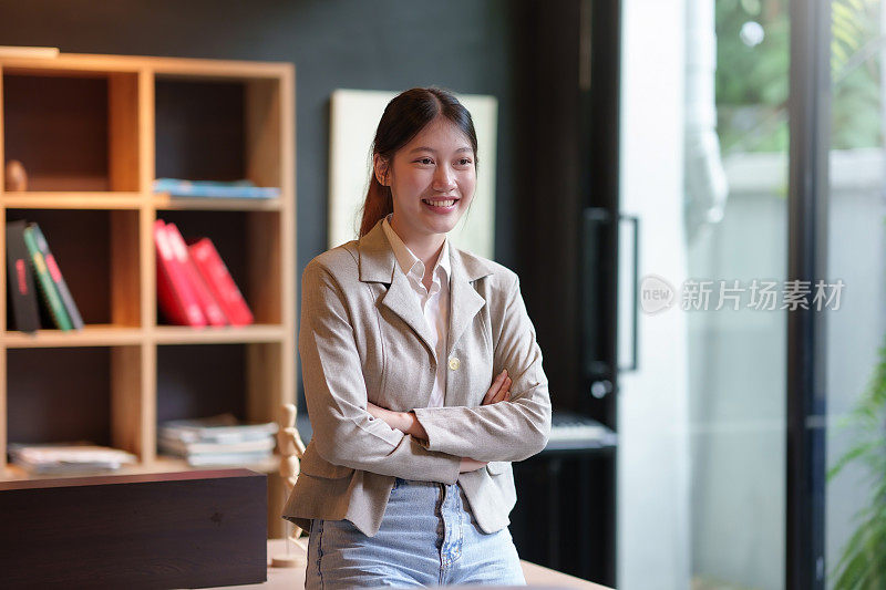 一个迷人的，美丽的亚洲女商人站在她的办公室里，双臂交叉，微笑着，开心地看着镜头。