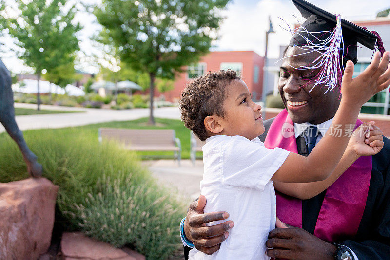 非裔美国男大学毕业生戴着帽子和长袍，在毕业典礼上和他三岁的儿子玩