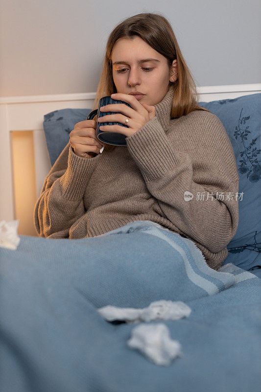 可爱的女孩穿着米色的毛衣，坐在床上，盖着蓝色的毯子，喝着热饮。