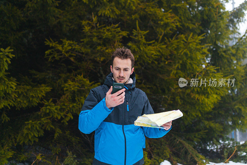 年轻的男性徒步旅行者穿着蓝色夹克，在雾蒙蒙的雪山中迷失了方向，一边看地图，一边看智能手机