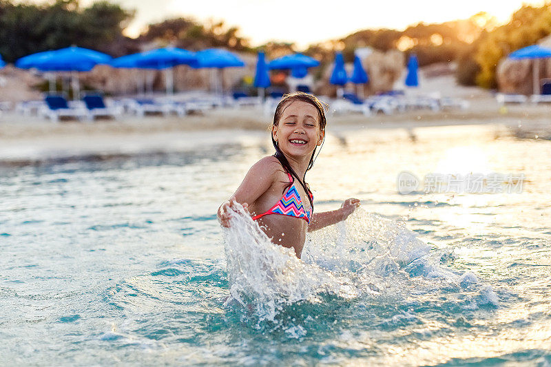 一个年轻可爱开朗的女孩穿着明亮的泳装在海里，玩耍，跳跃，享受游泳和飞溅的水在阳光的光芒在背光下在夏天的假期，在海滩上，文字的空间