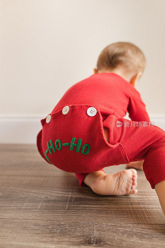 一个11个月大的圣诞宝宝穿着红色复古风格的圣诞睡衣，底部写着“HO-HO-HO”，在木地板上玩耍，为他的生日和2022年的假期做准备