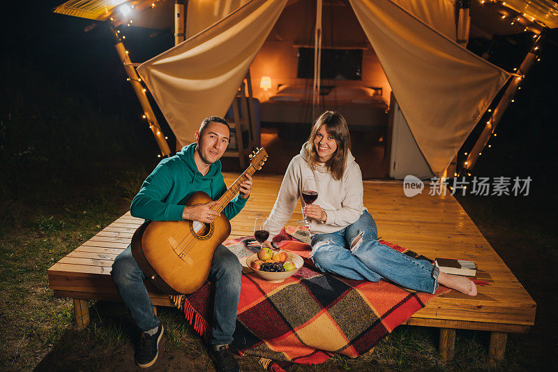 幸福的情侣在秋天的晚上豪华露营，在舒适的篝火旁喝着酒，弹着吉他。户外休闲休闲的豪华露营帐篷。生活方式的概念