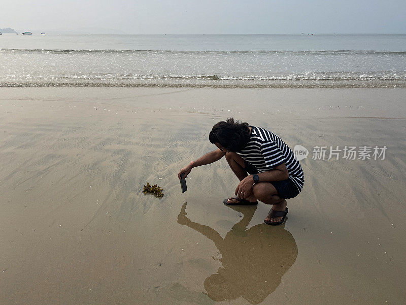 印度果阿邦帕洛勒姆，一个印度人在果阿海滩度假，蹲着用智能手机拍摄海水边沙滩上的海藻，度假游客在潮湿的海滩上反思，这是前景的焦点