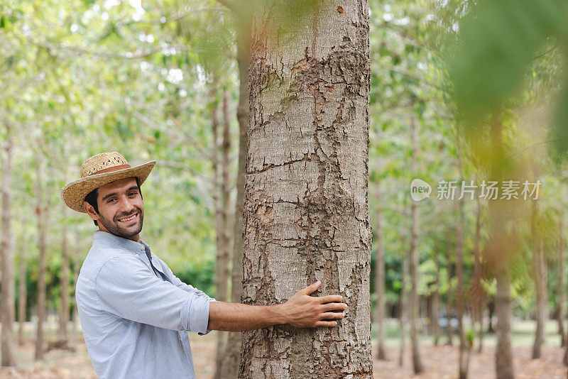 穿着白衬衫留着胡子的英俊男子倚在一棵树上，对着镜头微笑。