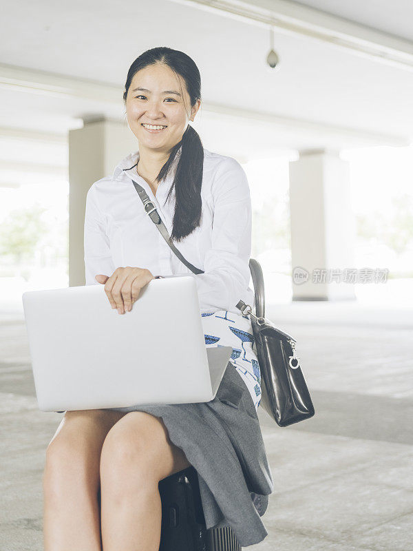 亚洲女商人坐在行李上，在机场使用笔记本电脑