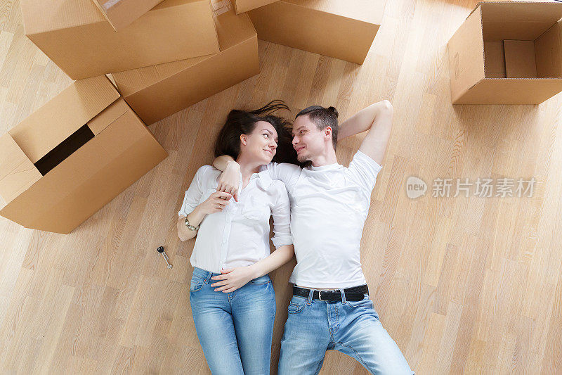 一对年轻夫妇躺在他们新房子的地板上，他们周围有许多包装纸箱。俯视图