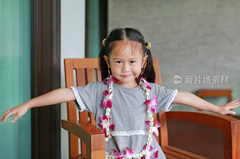 微笑的小亚洲女孩的肖像与欢迎兰花花花环坐在椅子上看相机。