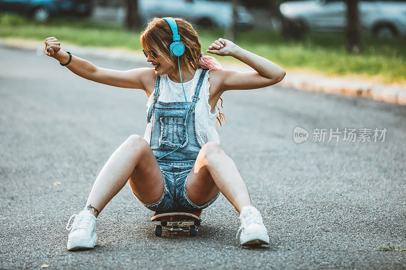 年轻女子戴着耳机坐在路上的滑板上。在一个阳光明媚的日子里，在公园里玩滑板的漂亮年轻女子。年轻迷人的美丽和时尚的年轻女人与滑板摆姿势的照片。她站在C里享受