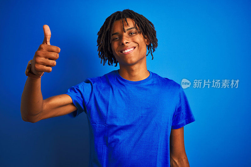 美国黑人男子与雷鬼发穿t恤站在孤立的蓝色背景做快乐的拇指手势与手。赞许的表情看着镜头，表示成功。