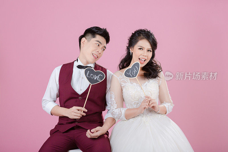 幸福的年轻新娘和新郎拿着心形木标签在粉红色的背景。新婚夫妇，新家庭，婚纱。新娘的婚礼。爱的概念