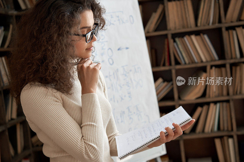 年轻的拉丁数学学校女教师戴着眼镜拿着笔记本思考课堂上的任务。西班牙语大学学院导师，研究生在校课堂教学。