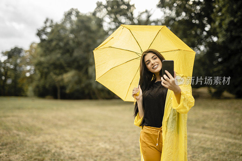 美丽的女人穿着黄色雨衣，拿着黄色雨伞，用手机自拍，享受雨天
