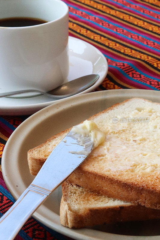 特写图片:两片涂了黄油的早餐，白盘子上的白吐司，黄油刀，黑咖啡的杯子和碟子，茶匙，桌布，重点放在前景