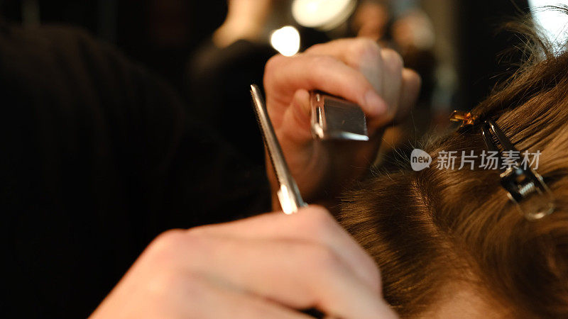 理发师用剪薄剪刀和金属梳子梳年轻人棕色的波浪状头发。在一个现代理发店在一个黑暗的关键闪电与温暖的光侧视图理发师服务