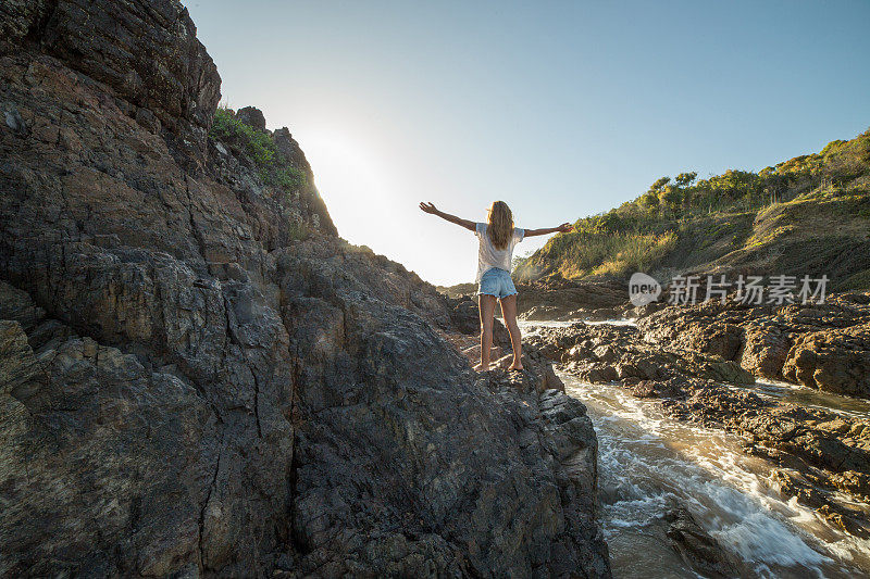 日出时，年轻女子在陡峭的岩石上张开双臂