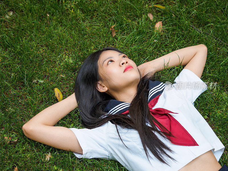 亚洲女学生的肖像在校服日本风格，俯视图十几岁的女孩躺在她的背部在夏季草地上，双手交叉在头下，白日做梦。