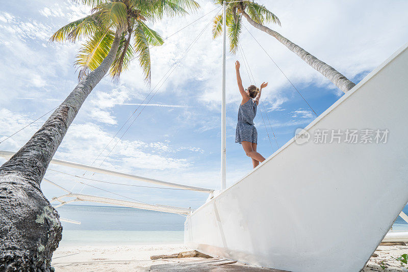 年轻女子在渔船上伸开双臂，棕榈树和热带海滩景观