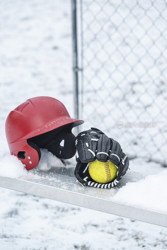 一个戴着垒球手套和棒球头盔的垒球坐在雪地里的长椅上