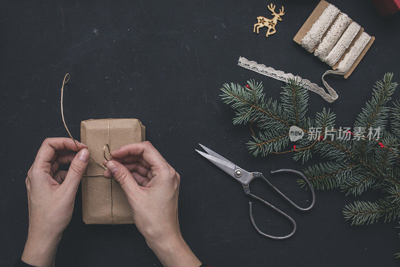 包装圣诞礼物盒。顶部视图的手在黑色的背景与冷杉树枝，装饰