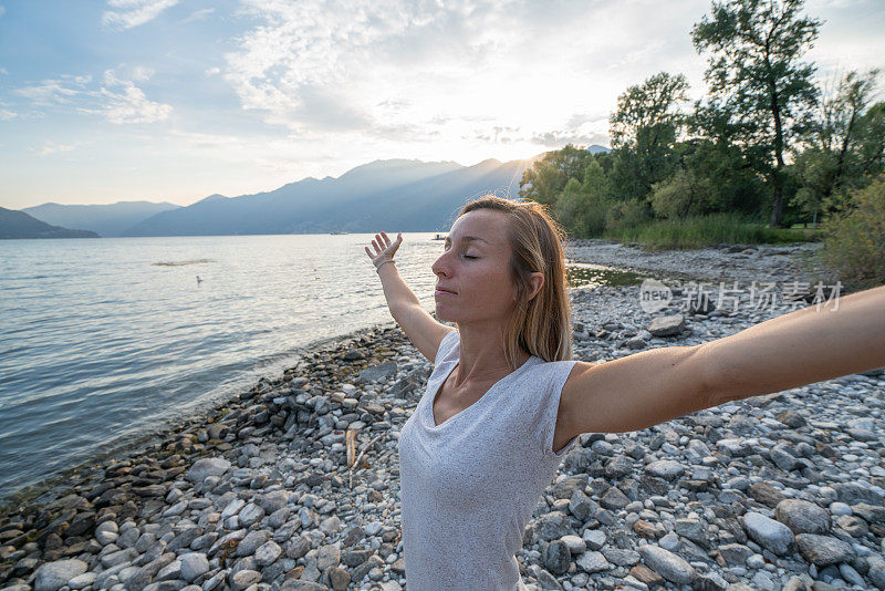 夕阳西下，拥抱大自然的快乐年轻女子;女性站在湖岸，以高山为背景——瑞士
