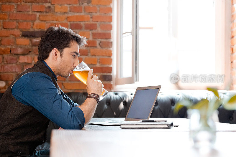 在酒吧里一边喝啤酒一边看笔记本电脑的男人
